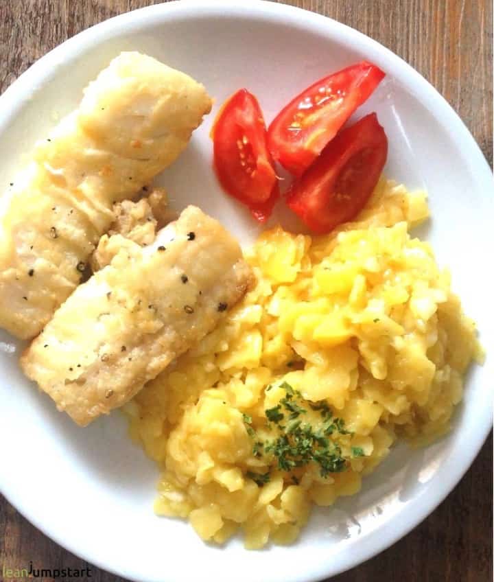 pan-fried cod fish fillets on a white plate with German potato salad and three tomato slices as garnish