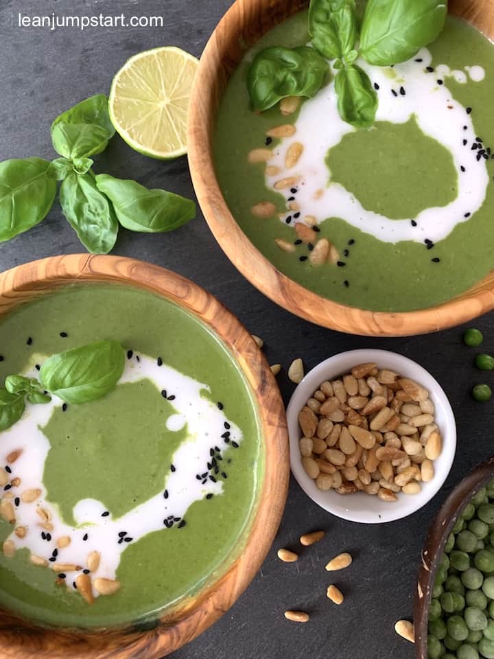 2 bowls of vegan broccoli soup with garnish surrounded by a lemon, and a small cup of pine nuts